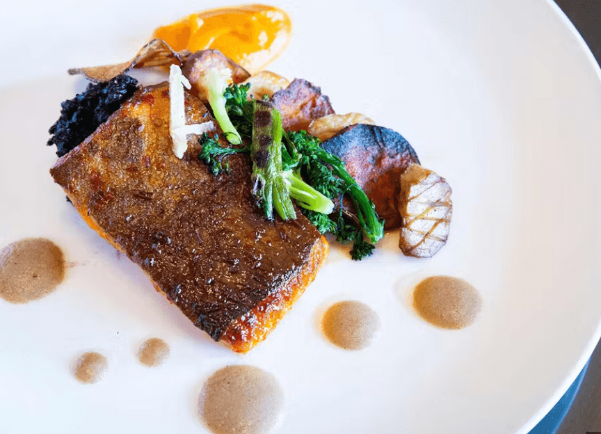 A gourmet dish inspired by the Culinary Institute, featuring crispy-skinned fish, roasted potatoes, greens, a bright orange sauce, delicate vegetable crisps, and artful brown sauce dots on a white plate.